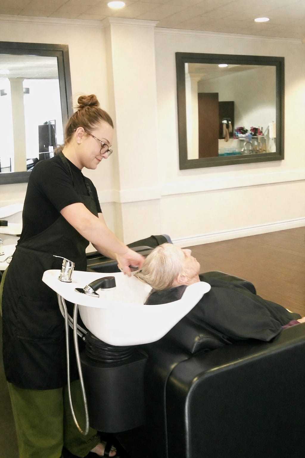 Stylist washing an elderly woman's hair in a salon sink.