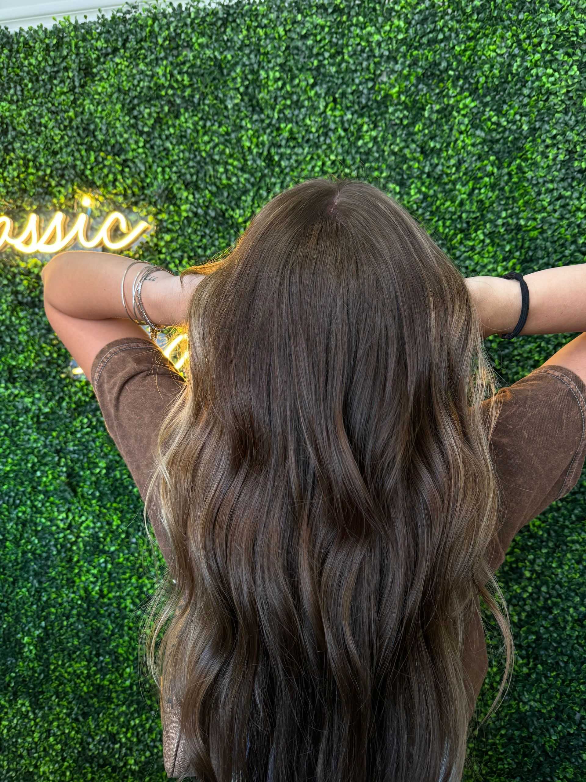 Woman with long hair stands against a leafy background with neon sign.